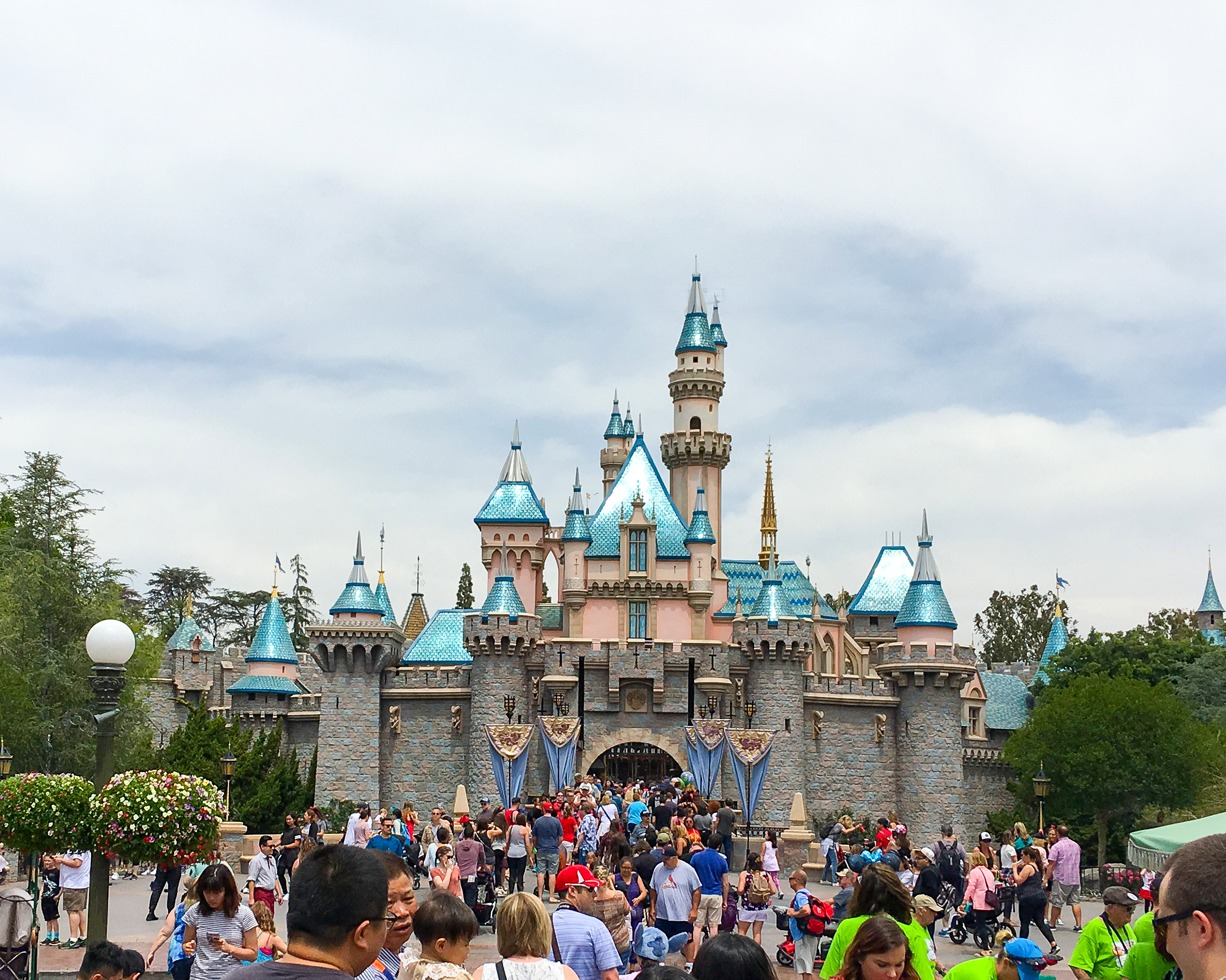 Sleeping Beauty Castle at Disneyland Park beneath a cloudy sky, with guests walking through the central plaza